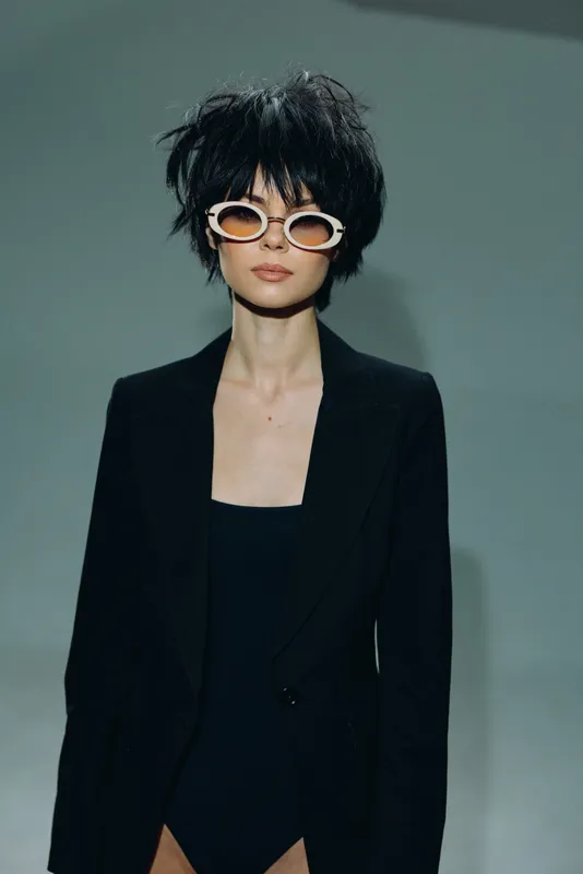 Stylish woman with short black hair wearing white sunglasses and a black blazer against a dark background