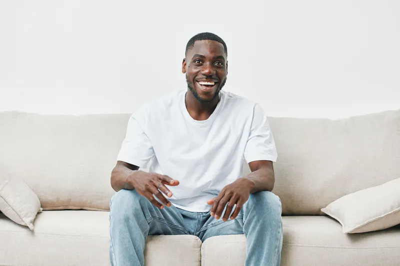 Happy young Black man in casual outfit smiling on a beige sofa, conveying joy and relaxation in a cozy indoor setting
