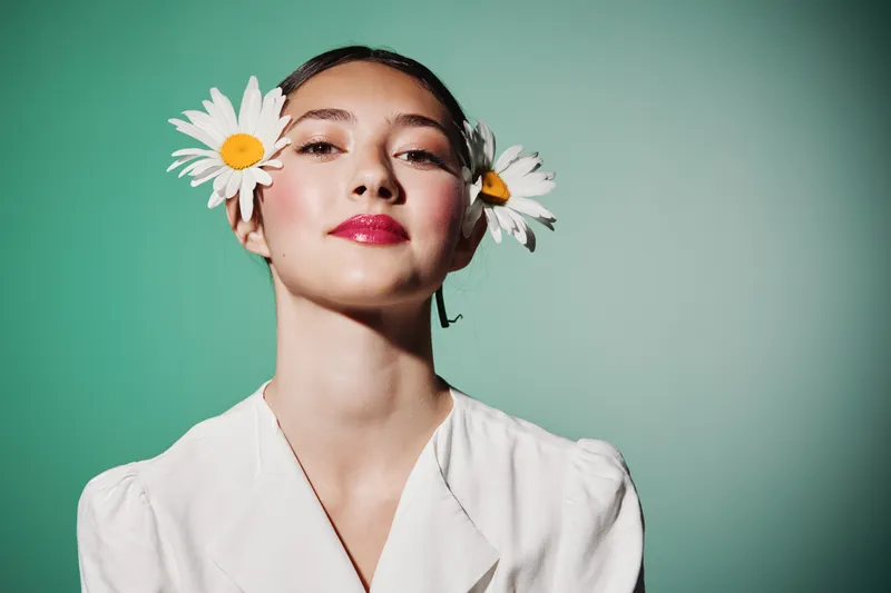 Young woman with natural makeup and bright red lipstick wears white flowers in hair. Her confident face and white blouse stand out against the vibrant green. Fashion beauty concept.