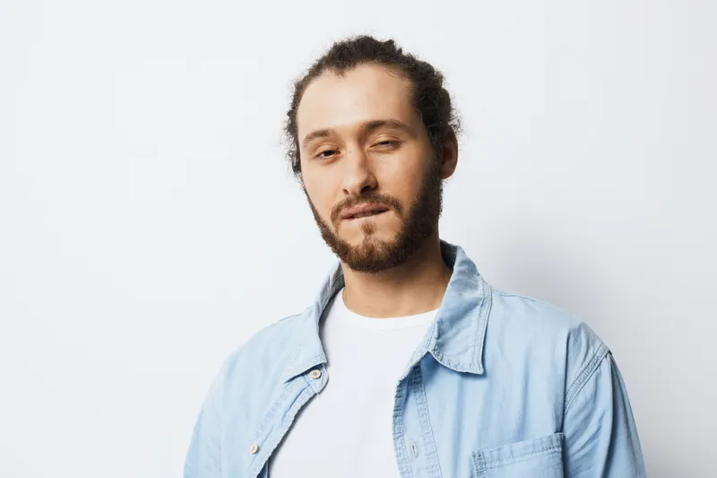 Confident young man with curly hair and beard wearing a casual denim shirt against a minimalistic white background, showcasing a playful yet relaxed expression
