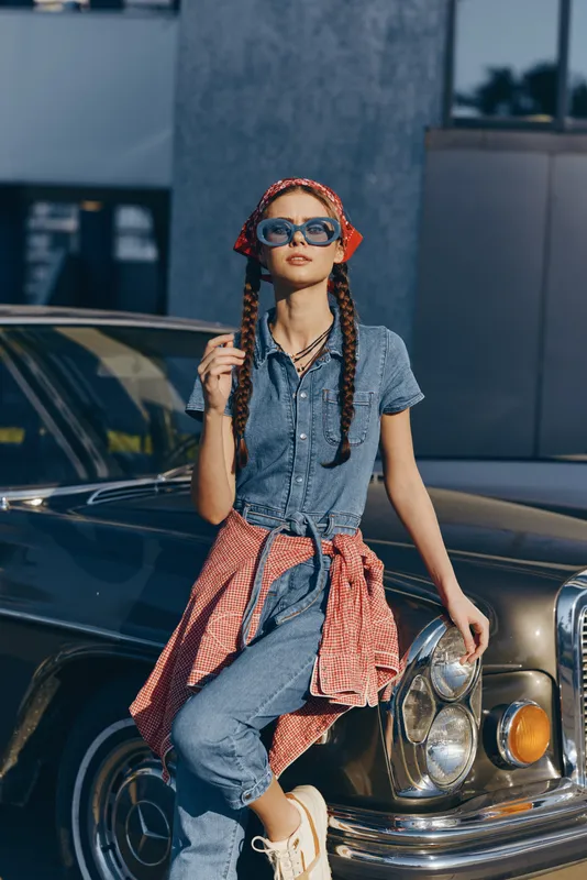 Lifestyle woman in modern boho-western denim street style with sunglasses and braided hair, posing confidently by a vintage car in film color tones.