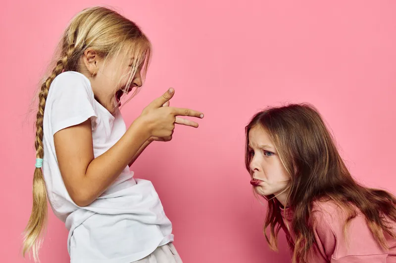 Two young girls interact on a pink background. One girl with a braid playfully points fingers like a gun while the other girl looks upset with a pout. Concept of childhood emotions and play.