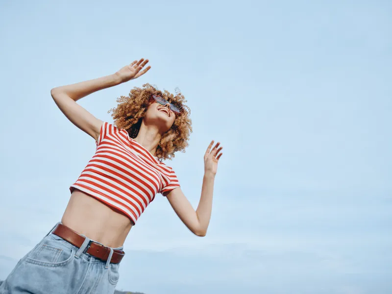 Young woman with curly hair wearing a red and white striped crop top and jeans, posing carefree against a bright blue sky, capturing playful movement and energy.