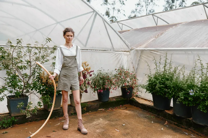 Young woman in green overalls holding a basket, exploring a greenhouse filled with blooming flowers, radiating joy and connection to nature