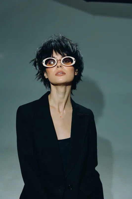 Stylish woman with short hair wearing white sunglasses and a black blazer posing confidently in a minimalist setting.