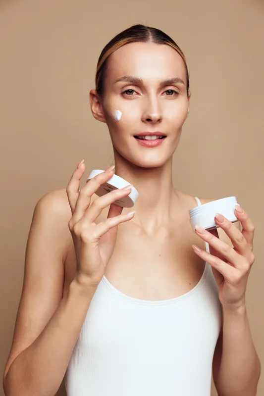Woman with clear skin applying face cream with fingers, skincare routine, natural beauty, neutral background, soft lighting, minimal makeup, healthy lifestyle, closeup portrait