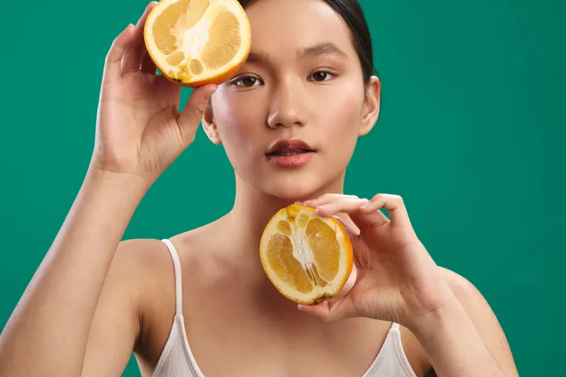 an asian woman holds sliced citrus fruits near her face in a bright studio setting, creating a fresh and playful portrait that highlights vitality, natural beauty, and a vibrant, clean mood.