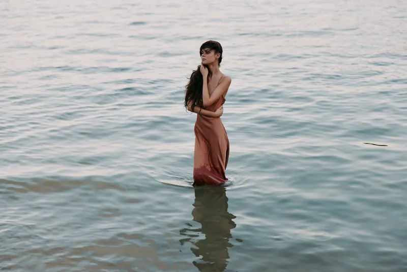 A lone woman stands waist-deep in calm water, wearing a flowing dress that drapes softly. The scene conveys calm reflection, graceful posture, and a tranquil coastal mood.