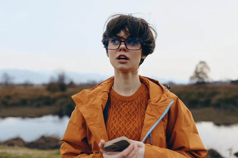 Woman lifestyle outdoors, wearing orange jacket and glasses, standing by a river bank, holding smartphone and looking ahead, calm autumn mood, natural light, shallow depth