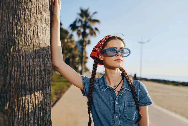 lifestyle woman in modern boho-western denim street style with braided hair and blue sunglasses, standing by palm tree in warm film color lighting, relaxed mood