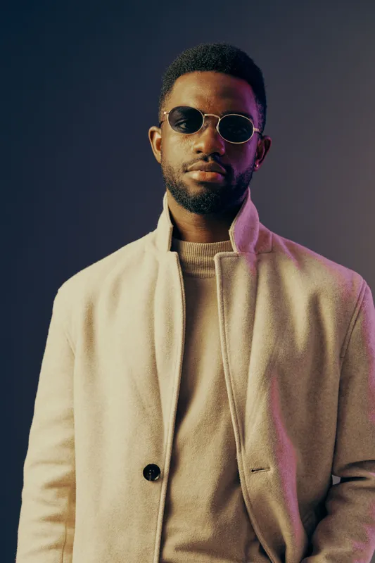 Stylish young African man wearing sunglasses and a beige coat against a purple blue gradient background, exuding confidence and modern fashion trends