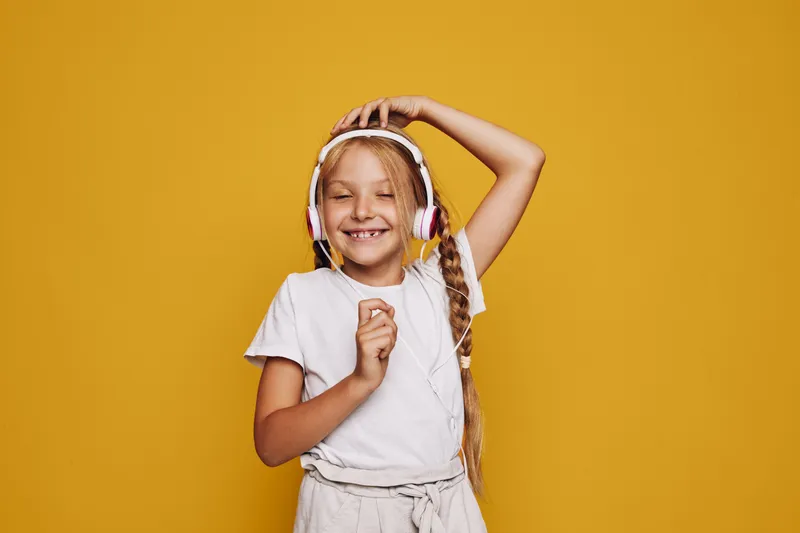Smiling girl with braided hair enjoys music on headphones with closed eyes on yellow background.