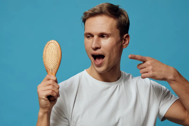 Young man with short brown hair, wearing a plain white t-shirt, holding a wooden hairbrush and pointing at his head with an expressive surprised look isolated on a bright blue background, studio shot.
