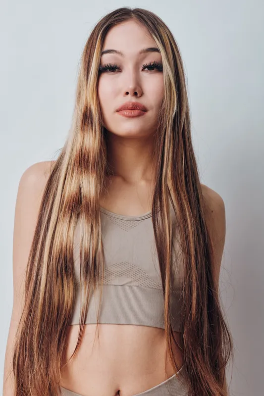 Young woman with long straight hair wearing casual top stands against plain background looking confidently at camera with neutral expression and natural makeup.