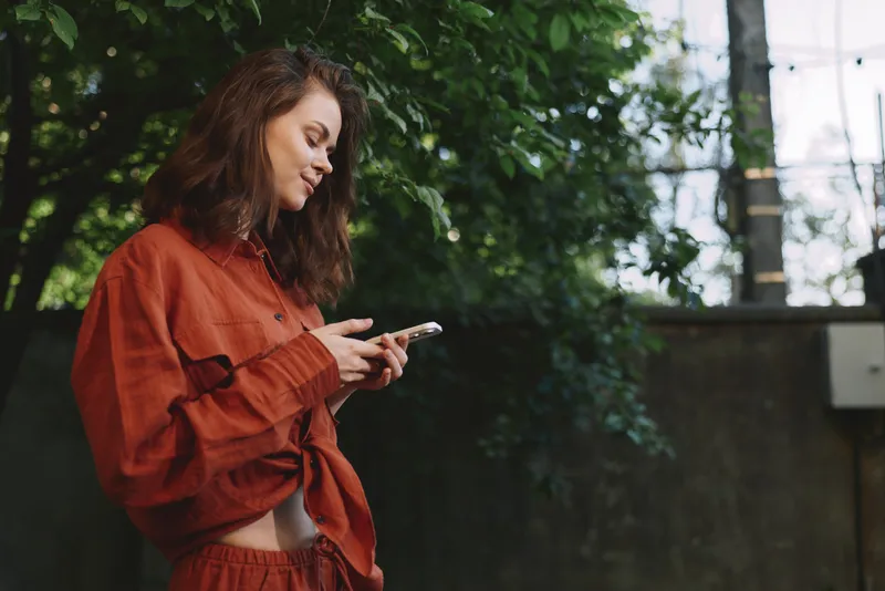 A woman in orange loungewear stands outdoors, focused on her phone among lush greenery. A candid lifestyle moment highlighting daily routine, connectivity, and relaxed urban living