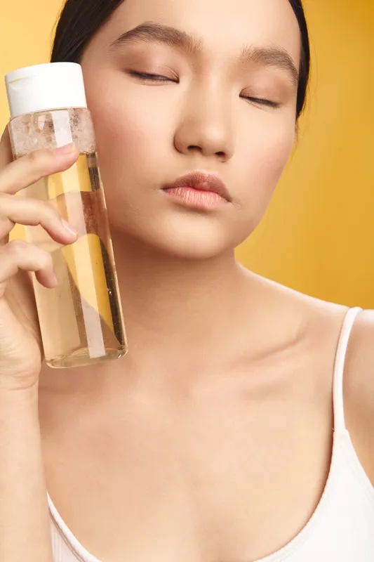Asian woman rests with eyes closed as she holds a clear bottle near her face, framed by a warm backdrop, evoking calm, self care, and a fresh everyday routine.