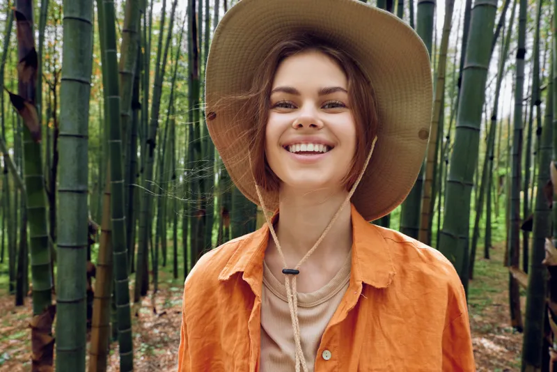 Woman smile portrait with hat in bamboo forest outdoors. Happy traveler wearing orange shirt, natural light and lush green stalks, leisure nature walk and eco travel mood.