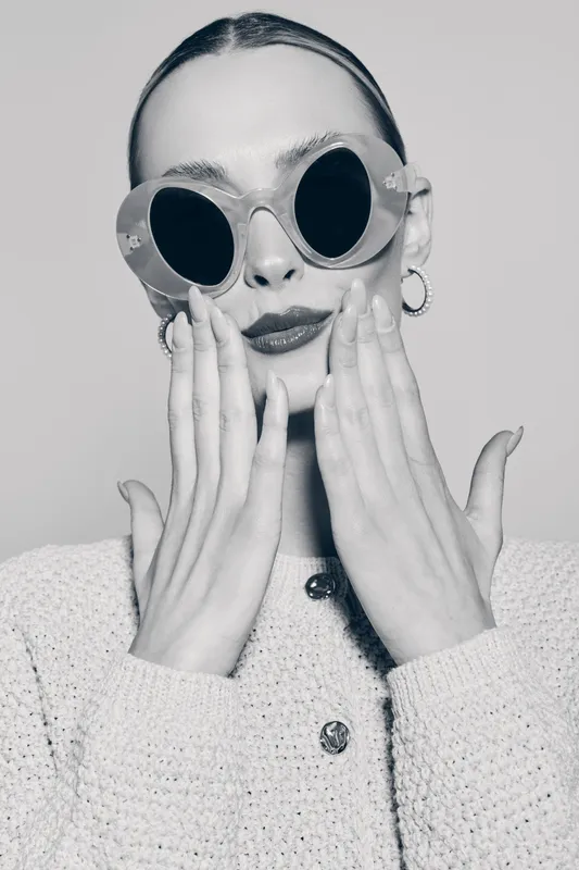 Portrait of a fair skinned Caucasian woman wearing large round sunglasses and knitted jacket touching her face in studio.