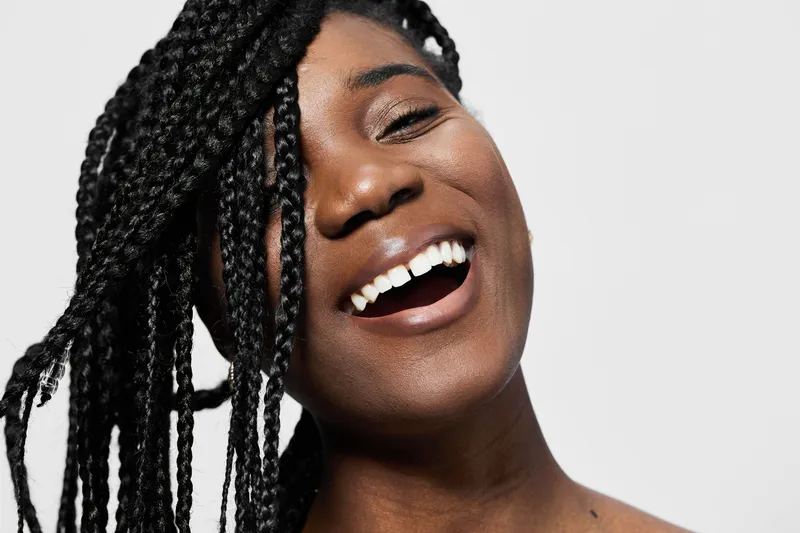 A joyful African woman with braided hair smiles brightly, showcasing her teeth against a clean, neutral background, perfect for lifestyle and beauty concepts