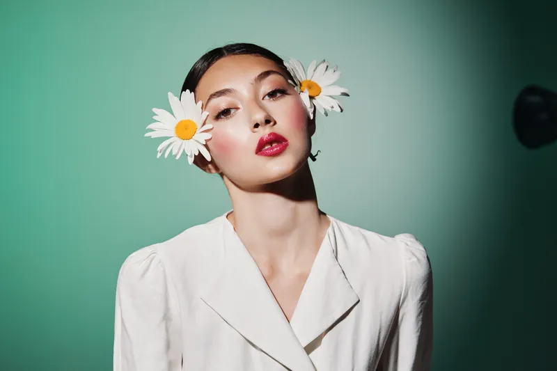Young woman in white fashion jacket wears fresh daisy flowers in her hair with bold glossy red lips and clean makeup. Studio shot on muted green background. Beauty creative fashion concept.