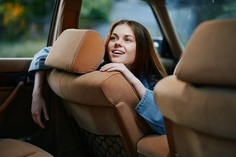 Happy young woman enjoying a road trip, leaning over the front seat in a cozy car with warm colors and soft lighting, reflecting a joyful travel experience.