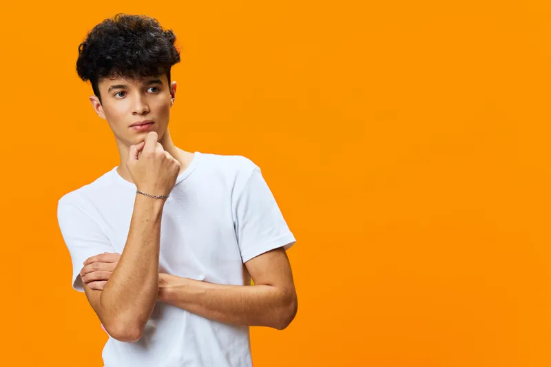 thoughtful young man with curly hair wearing a white t-shirt on vibrant orange background, posing with hand on chin, casual style, isolated studio portrait