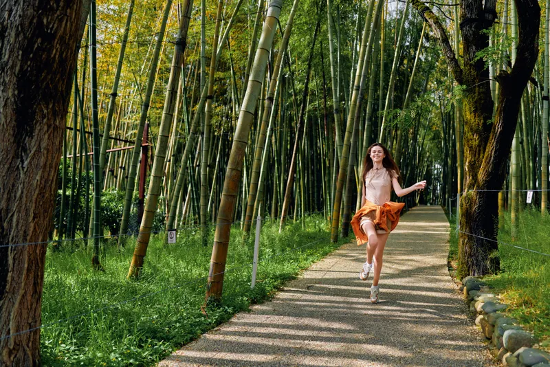 woman running bamboo forest path smiling as she leaps along a sunlit trail in casual summer dress, joyful motion and freedom, outdoor nature scene for leisure and travel