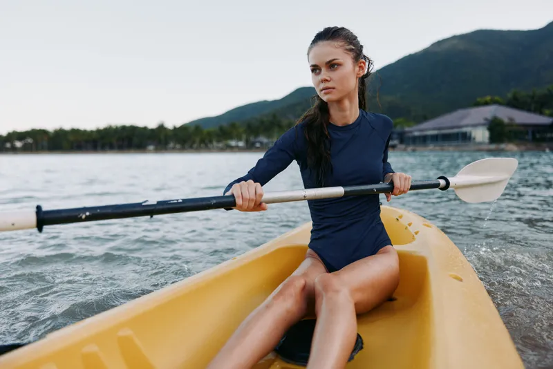 A focused kayaker in a blue swimsuit paddles a bright yellow kayak across calm water, with distant mountains and a clear sky creating a vivid outdoor sport scene.