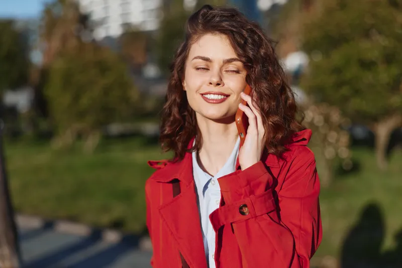 Young woman with curly brown hair wearing a red coat talks on a smartphone outdoors with a joyful expression on a sunny day in a park setting.