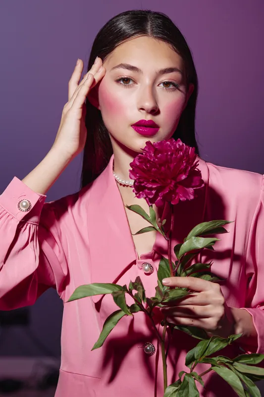 Young woman wearing a bubblegum pink jacket poses with vibrant fuchsia flower held near neck. Her bold makeup with purple lips and bright blush matches well. Creative beauty fashion concept.