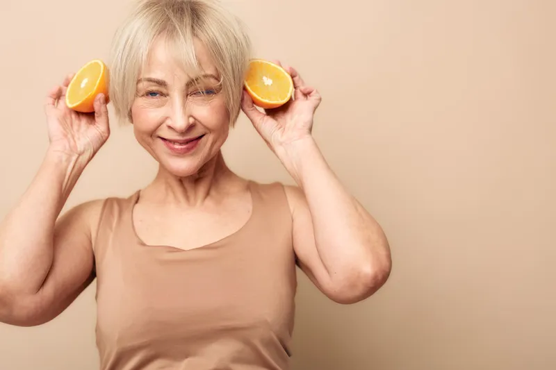 Smiling mature woman holds two orange halves near her ears, wearing beige sleeveless top on beige background. Concept of healthy lifestyle, natural beauty, wellness, and vitamin C benefits.