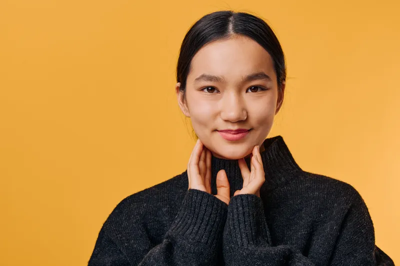 Portrait of young Asian woman with black hair wearing dark knit sweater standing against solid yellow background with hands near neck and smiling softly at camera.