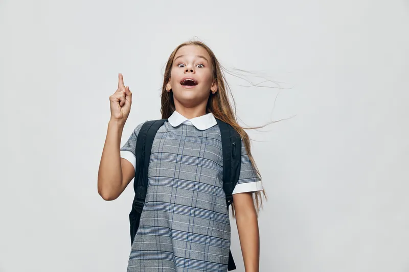 Girl with backpack in dress pointing upward with surprised face on gray background for education, school, learning and childhood concept with long hair and casual look.