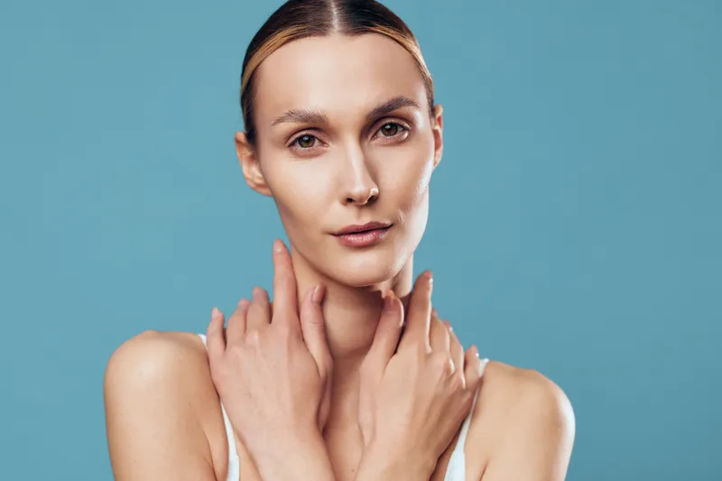 Young woman with clear skin and pulled back hair posing with crossed hands on neck, neutral expression, soft lighting, blue background, beauty and skincare concept