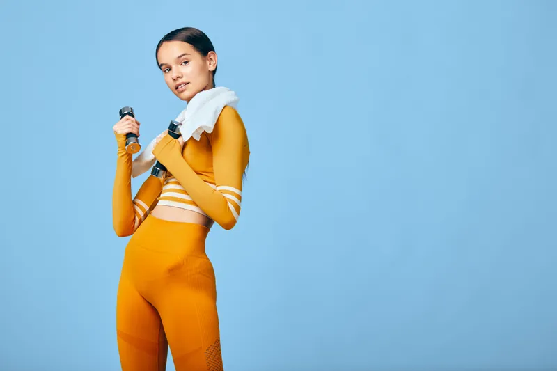 Woman fitness athlete in an orange workout outfit with a towel over her shoulder, holding a water bottle against a blue studio backdrop, showcasing sport, training, and energy.