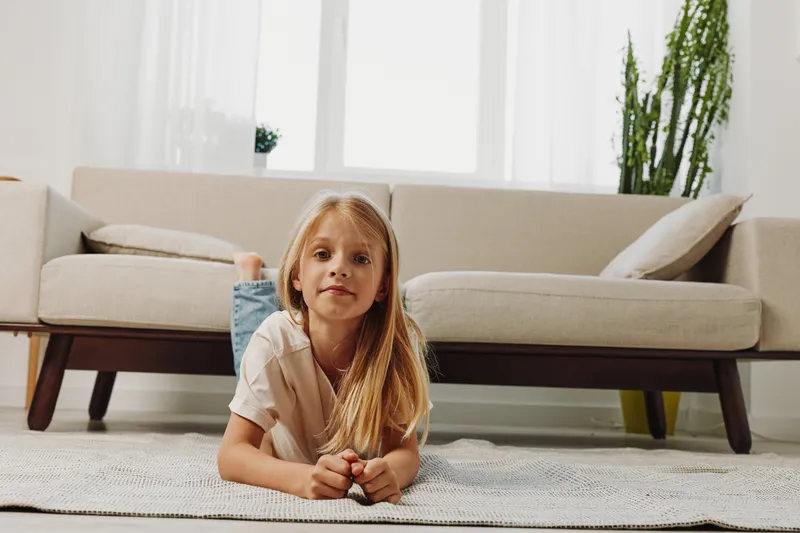 Blond girl lying on carpet in bright living room with modern sofa and natural light. Indoor home environment, child portrait, cozy family interior with plants and soft neutral colors.