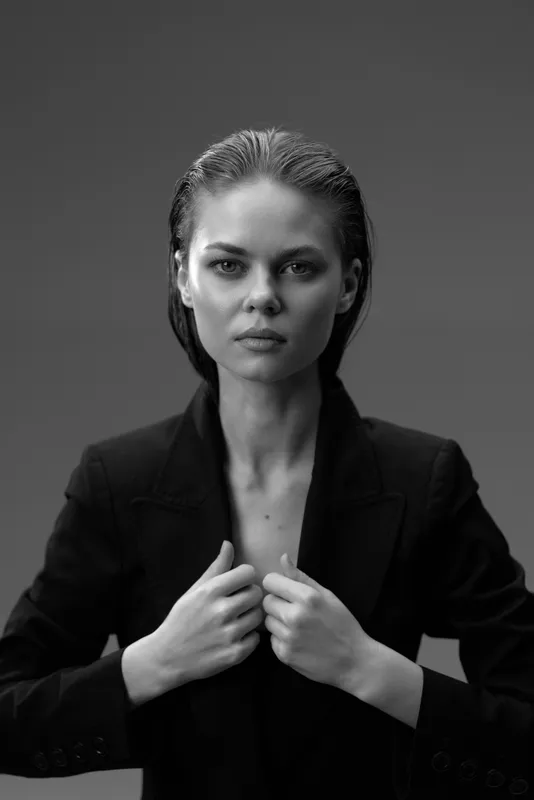 Black and white portrait of serious woman in black blazer with intense expression against dark background.