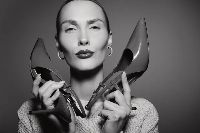 Elegant white woman with pale skin holds two high heel shoes close to her face, posing confidently in monochrome. Concept of fashion, style, and footwear elegance.