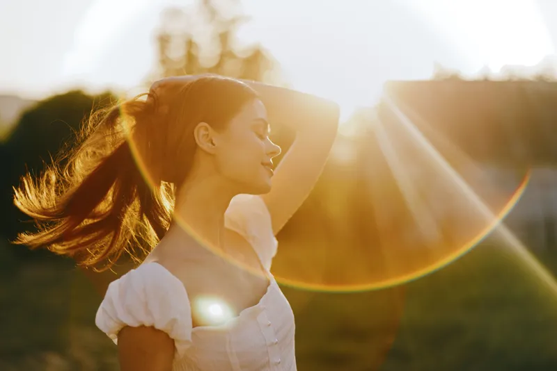 Young woman enjoying sunset outdoors, with flowing hair illuminated by golden sunlight, conveying joy and freedom in a serene natural setting
