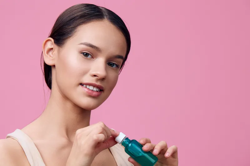 Young woman applying skincare serum, glowing skin, light pink background, healthy beauty concept, wellness routine, natural product, confidence
