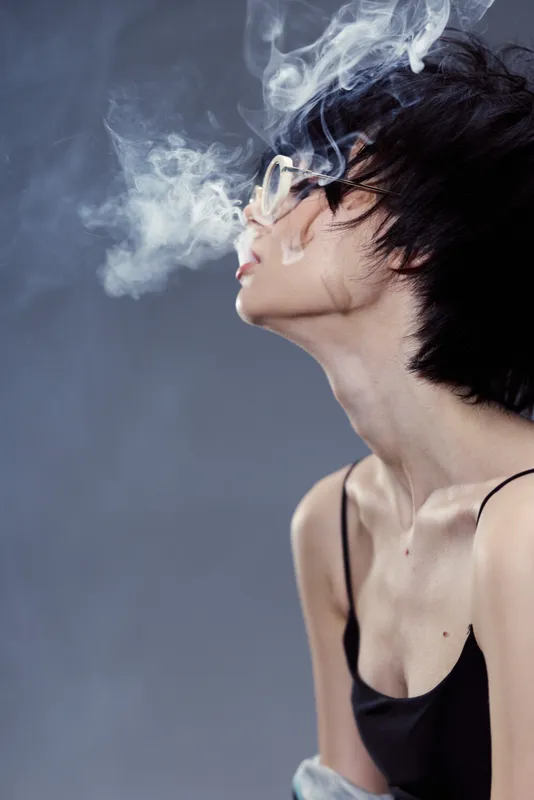 Young woman with short hair blowing smoke, wearing glasses and a black top with a moody dark background.