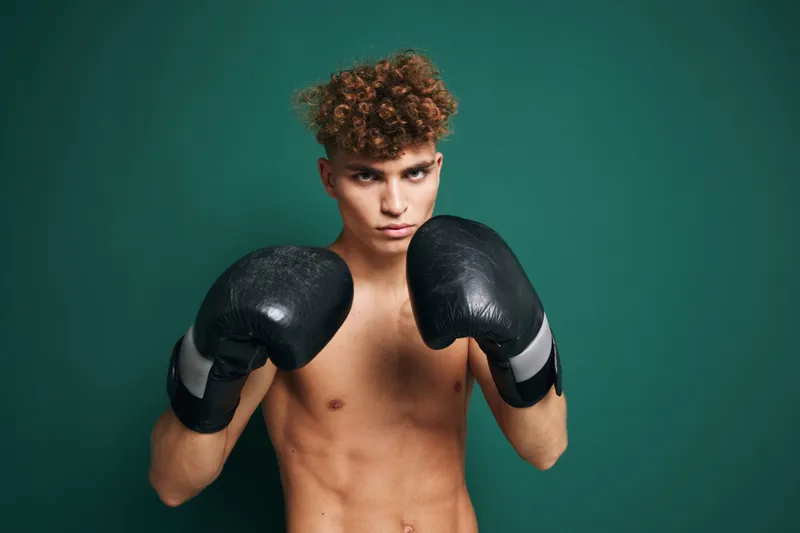 Young male boxer with curly hair wearing black boxing gloves in fighting stance against green background. Athlete training, fitness, strength and focus for combat sport and workout.