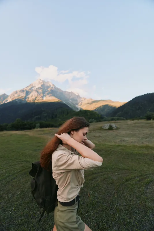 A young woman with a backpack walks along a grassy trail in a mountain valley, adjusting her hair as warm sun lights the snowcapped peaks.