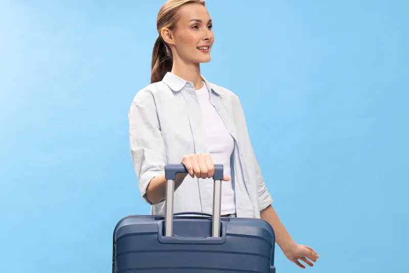Young woman with suitcase standing against blue background looking away with slight smile. Concept of travel, vacation, preparation, and lifestyle with casual outfit and relaxed mood.