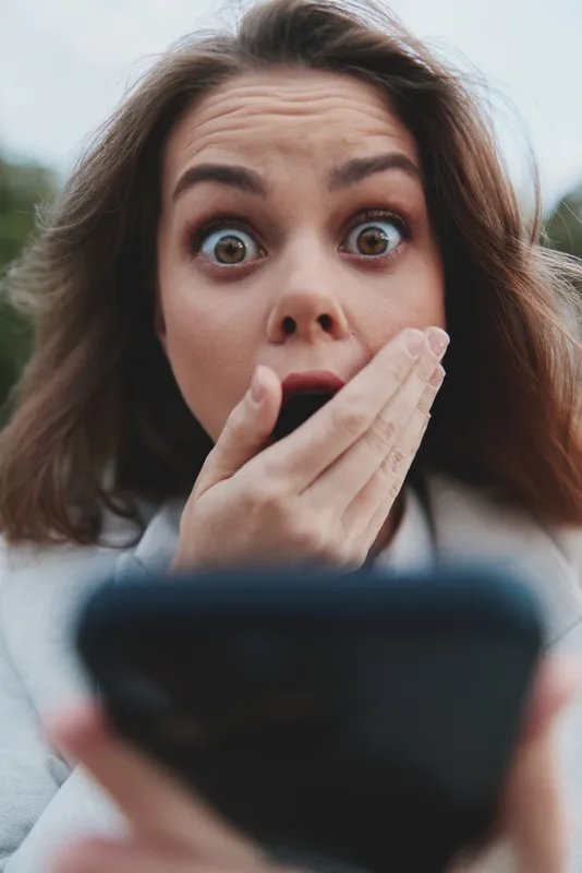Surprised businesswoman in casual attire reacting to news on her smartphone outdoors with a shocked expression, showcasing modern professional life.