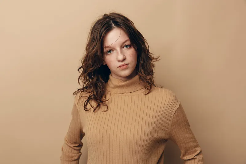 Young woman with curly brown hair wearing beige knitted sweater looks confidently at camera against plain beige background. Concept of natural beauty, casual style, and autumn fashion.
