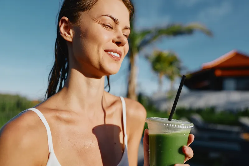 Cheerful woman outdoors enjoying a green smoothie, sunlit moment, relaxed posture, light breeze, natural colors, lush backdrop, healthy lifestyle, casual confidence, warm atmosphere