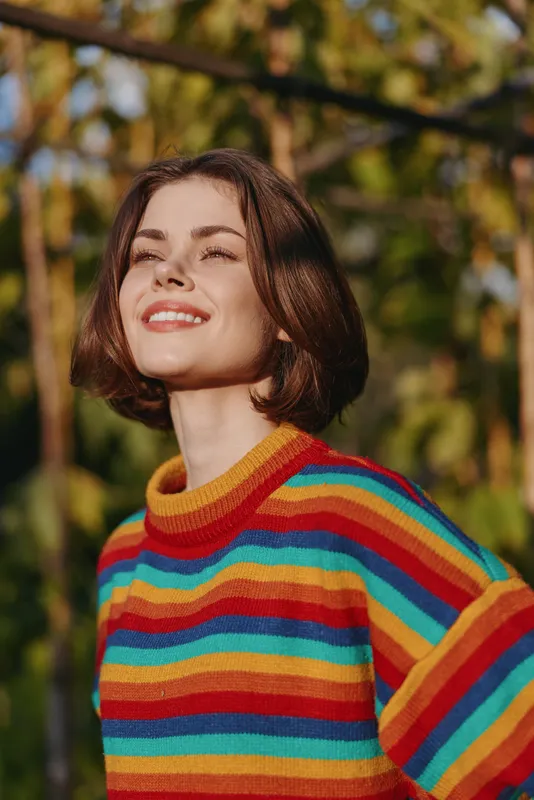 Woman portrait smile sweater outdoors sunlight happy casual young female in colorful striped knitwear, bob haircut looking up with serene joyful expression in park under warm light