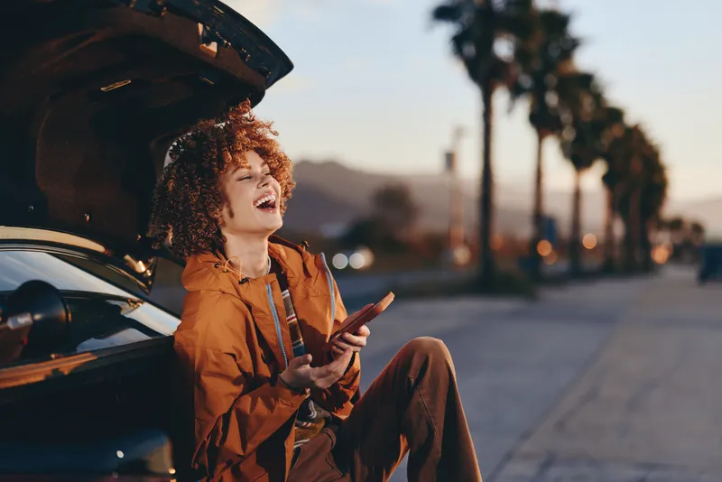 Woman lifestyle shows a woman in orange jacket with curly hair laughing holding a phone beside an open car hood outdoors at sunset on quiet roadside street with warm evening light