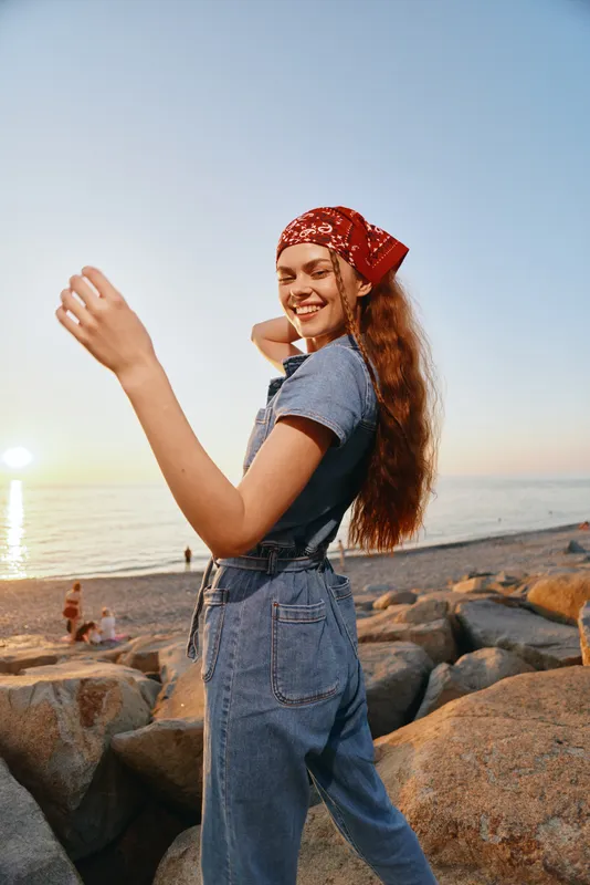 Lifestyle woman in modern boho-western denim street style smiling outdoors by the rocky beach at sunset, film color tones creating warm mood and natural light.